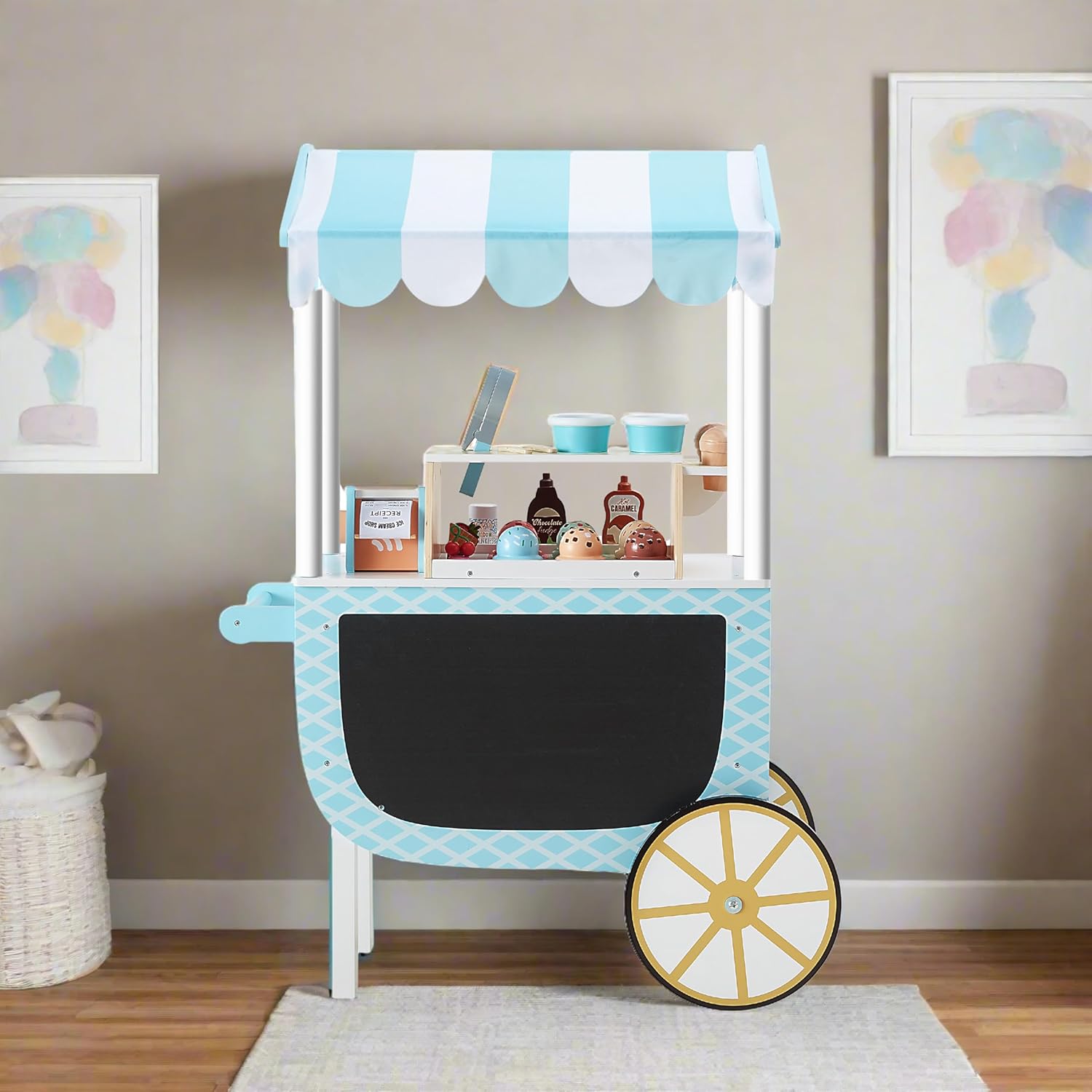 A blue and white toy ice cream cart with a striped canopy, shelves holding play ice cream items, and a large chalkboard panel stands in a room with pastel wall art.