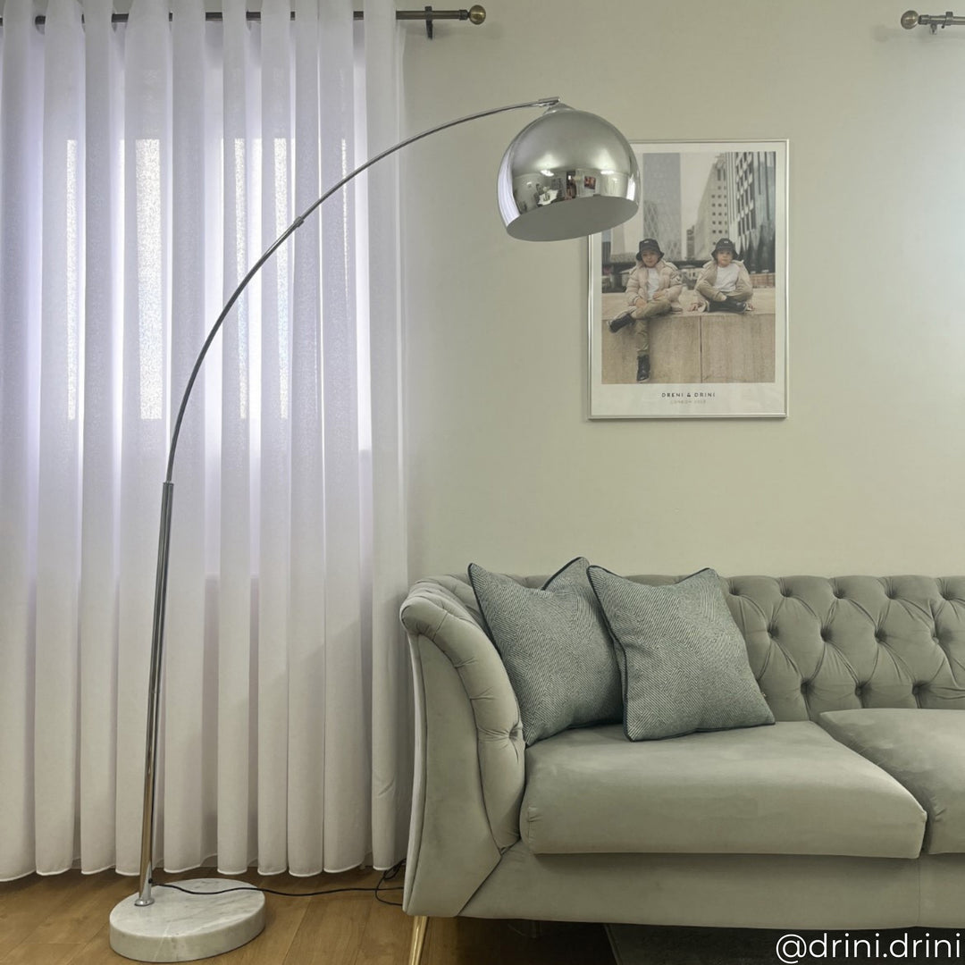 A modern living room features a tufted gray couch, a curved floor lamp with a metallic shade, and a framed wall photo. Light filters through sheer white curtains.