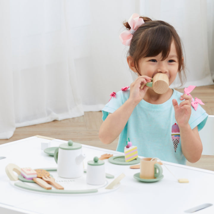 A young girl in a blue shirt with pink bows in her hair is pretending to drink from a toy cup at a white table set with toy tea items and pastries.