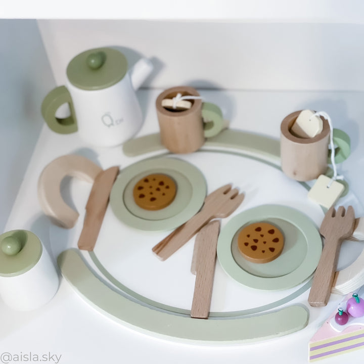 A children's wooden tea set from Teamson Kids laid out on a white surface, featuring a teapot, cups with tea bags, plates with cookies, and utensils in soft pastel colors.