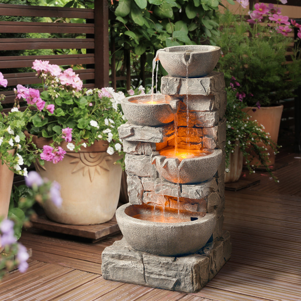 Gray water fountain with lights under water in each tier next to some potted plants
