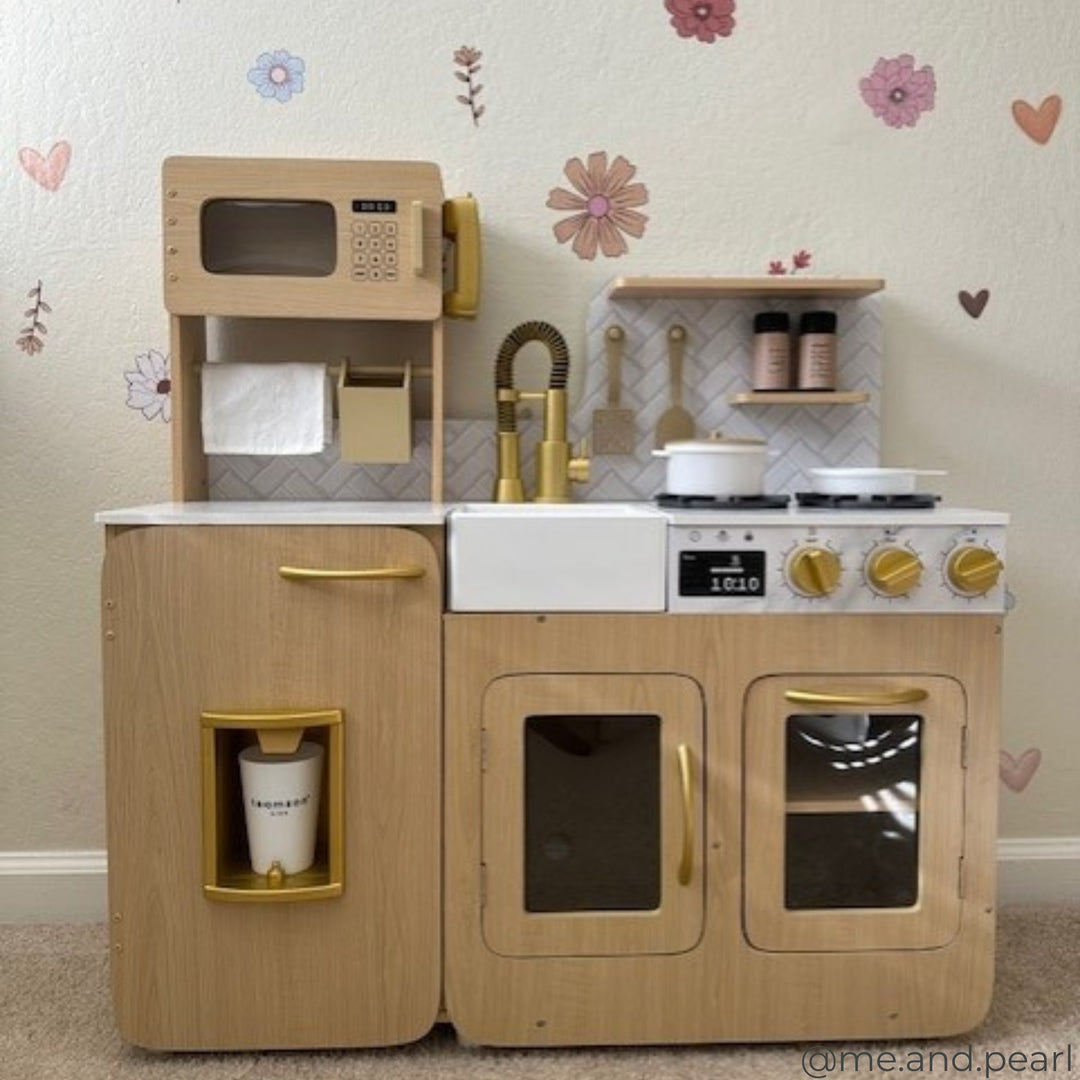 A wooden toy kitchen set with a stove, microwave, sink, and refrigerator. The kitchen has gold accents and is placed against a wall with flower decals.