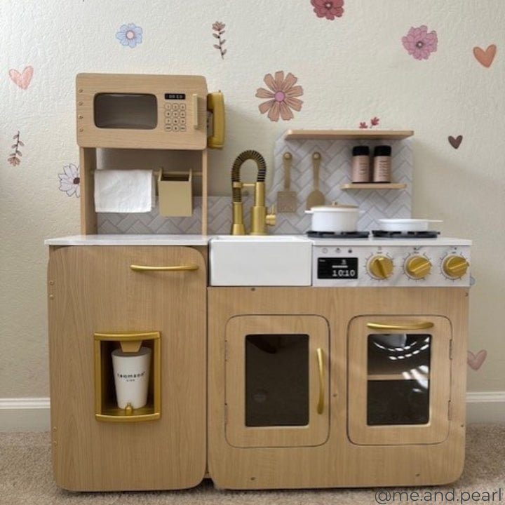 A wooden toy kitchen set with a stove, microwave, sink, and refrigerator. The kitchen has gold accents and is placed against a wall with flower decals.