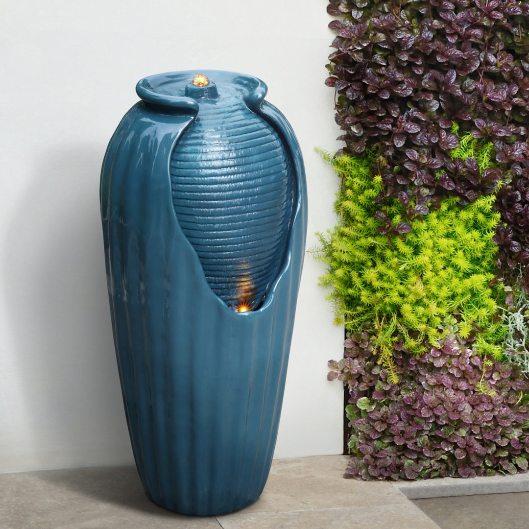 A tall blue faux vase water fountain with two lights next to some bushes